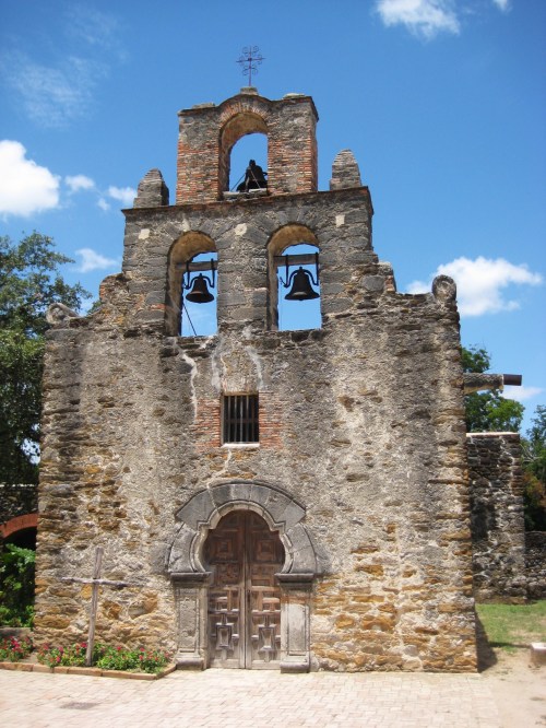 Historic stone church with a bell tower, featuring three bells and a wooden door, set against a blue sky.