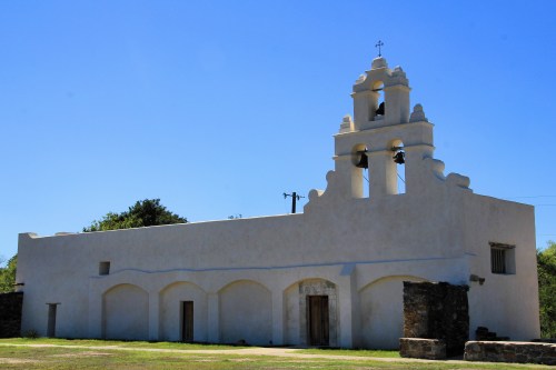 A white historic church with a bell tower against a clear blue sky, surrounded by greenery.