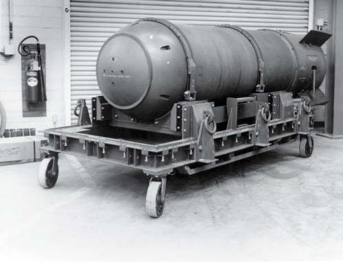 A large cylindrical object on a wheeled platform, likely a bomb or missile, in a warehouse setting.