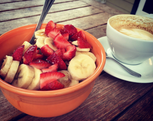 A bowl of sliced bananas and strawberries next to a cup of latte on a wooden table.
