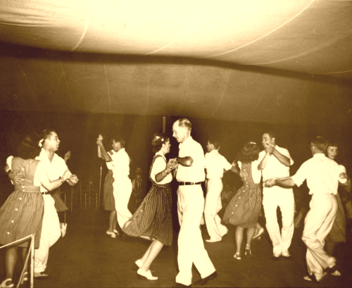 A vintage photo of couples dancing joyfully in a dimly lit hall, dressed in 1940s attire.