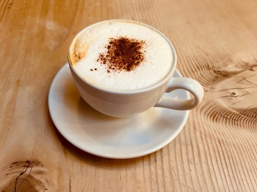 A white cup of cappuccino topped with a sprinkle of cocoa powder, resting on a white saucer on a wooden table.