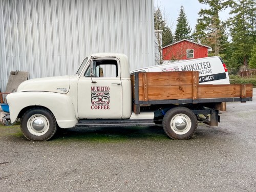 A vintage white pickup truck with a wooden bed, featuring a sign for Mukilteo Coffee on the side.