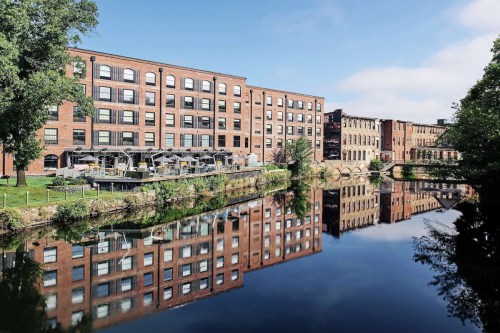 A brick building along a river, reflecting in the calm water, with outdoor seating and greenery nearby.