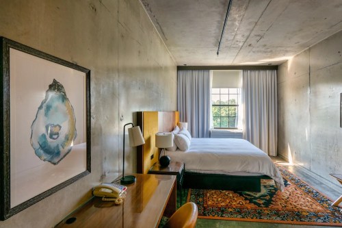 Modern hotel room with concrete walls, a bed, desk, and large window with natural light. Artwork hangs on the wall.