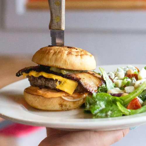 A hand holding a plate with a cheeseburger topped with bacon and a knife, alongside a fresh salad.