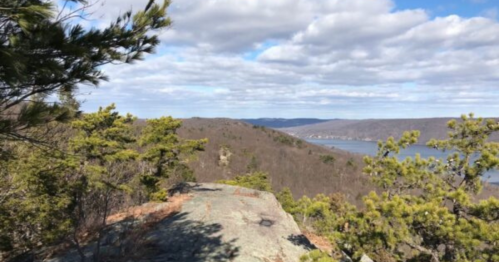 Scenic view from a rocky overlook, featuring rolling hills, trees, and a river under a partly cloudy sky.