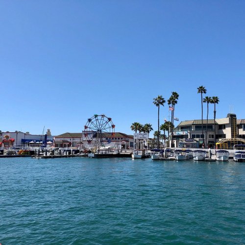 A vibrant waterfront scene featuring a ferris wheel, palm trees, and boats along a clear blue sky.