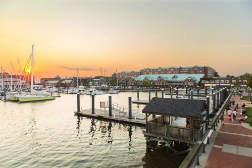 Sunset over a marina with boats docked, a waterfront promenade, and buildings in the background.