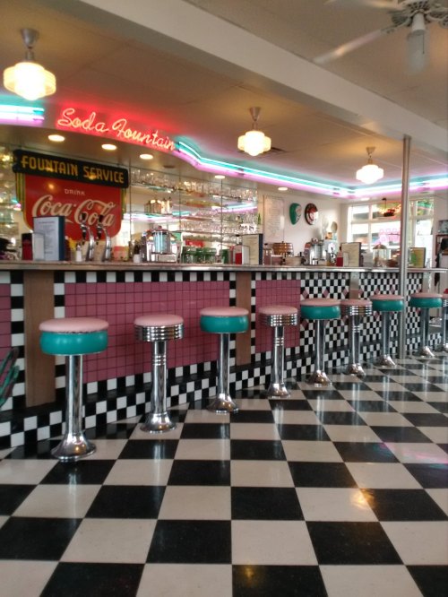 A retro diner interior featuring a checkered floor, colorful stools, and a neon "Soda Fountain" sign.