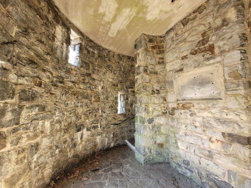 Interior of a stone structure with curved walls, small windows, and a stone staircase. Natural light filters in.