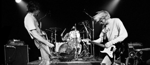 Black and white photo of a band performing on stage, featuring a guitarist and a drummer in action.