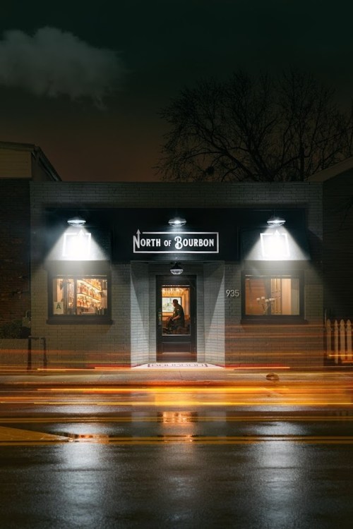 A dimly lit bar entrance, "North of Bourbon," with glowing lights and a person standing inside on a rainy night.