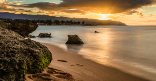 A serene beach at sunset, with smooth waves, rocky formations, and a colorful sky reflecting on the water.