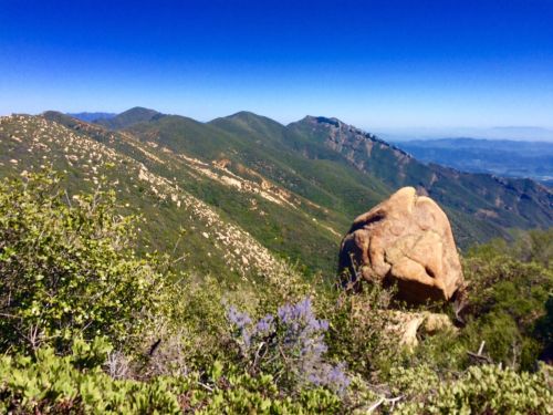 A scenic view of rolling green mountains under a clear blue sky, with a large rock formation in the foreground.