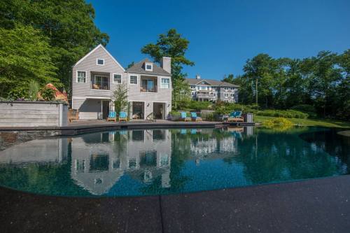 A modern house with a pool reflects in the water, surrounded by greenery and blue skies.