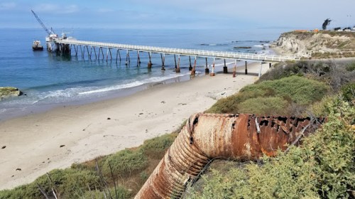 A rusty pipe leads to a sandy beach with a pier extending into the calm ocean under a cloudy sky.
