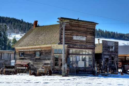 A rustic wooden building in a snowy landscape, featuring old signage and vintage equipment outside.