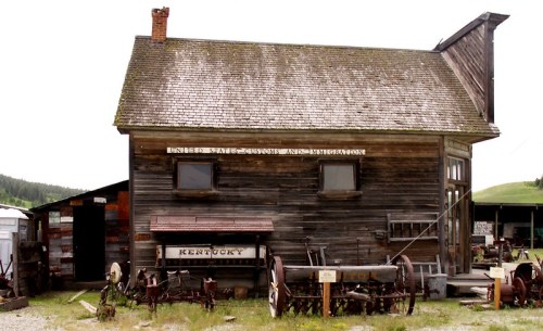 A rustic wooden building with a sloped roof, surrounded by old tractors and farm equipment, set in a grassy area.