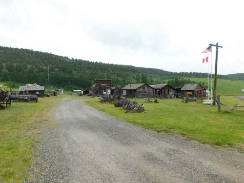 A dirt road leads through a rustic ghost town with wooden buildings and old machinery, surrounded by green hills.