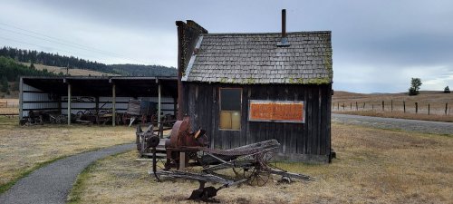 A weathered wooden cabin with a shingled roof, surrounded by dry grass and old farm equipment under a cloudy sky.