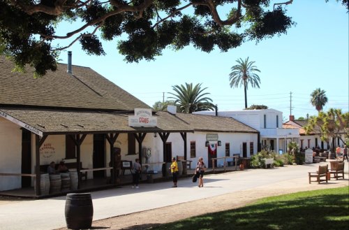 Historic buildings line a sunny street, with palm trees and people enjoying the outdoor space.