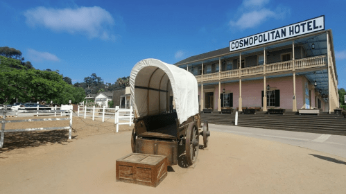 A covered wagon in front of the historic Cosmopolitan Hotel, surrounded by a dirt area and greenery.