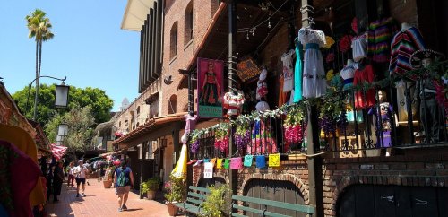 Colorful storefronts line a vibrant street, adorned with traditional clothing and decorations, with people strolling by.