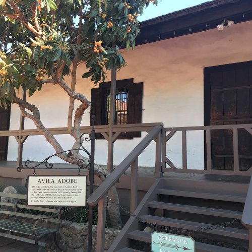 Historic Avila Adobe in Los Angeles, featuring a tree and entrance steps, with a sign detailing its history.