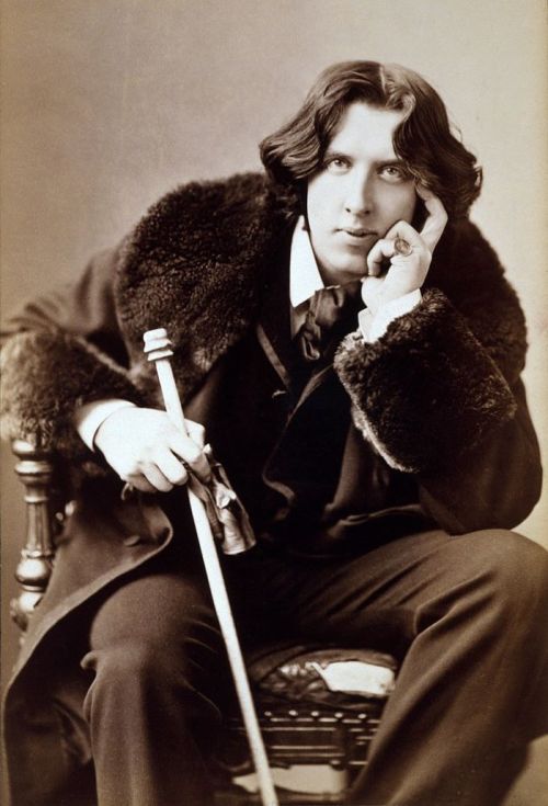 A sepia-toned portrait of a man with wavy hair, seated with a cane, wearing a fur-collared coat and a thoughtful expression.