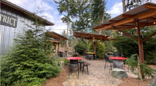 Outdoor dining area with red tables, surrounded by trees and a rustic building under a blue sky.