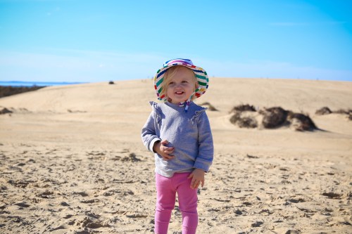 A smiling toddler in a colorful hat stands on a sandy beach, with dunes and a blue sky in the background.