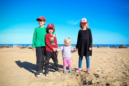 Four children stand on a sandy beach, smiling under a clear blue sky, wearing colorful hats and casual clothing.