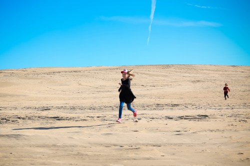 A girl runs joyfully on a sandy beach under a clear blue sky, with another person in the background.