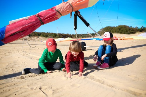 Three children play in the sand on a beach, with colorful kites and a clear blue sky in the background.
