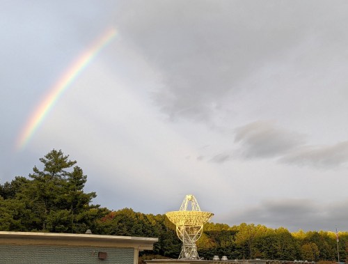 A rainbow arcs over a large satellite dish surrounded by trees and a cloudy sky.