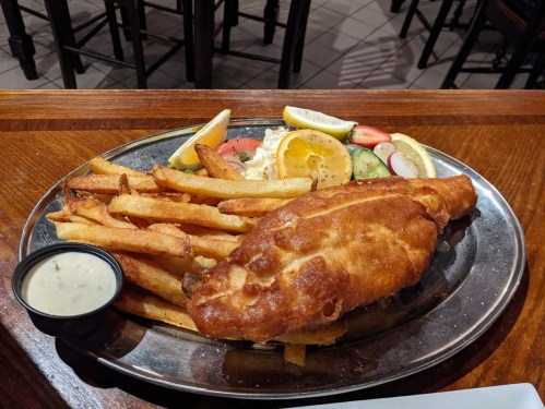 A plate of golden fried fish with crispy fries, lemon wedges, and a side of tartar sauce and fresh vegetables.
