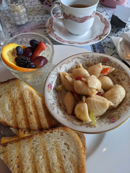 A plate with pasta salad, toast, and a glass of mixed fruit beside a cup of tea on a lace tablecloth.