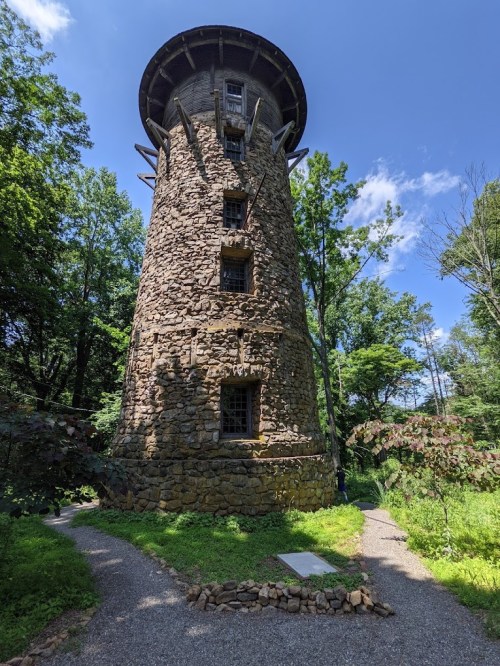 A tall stone tower surrounded by trees, with a clear blue sky above and a path leading to it.
