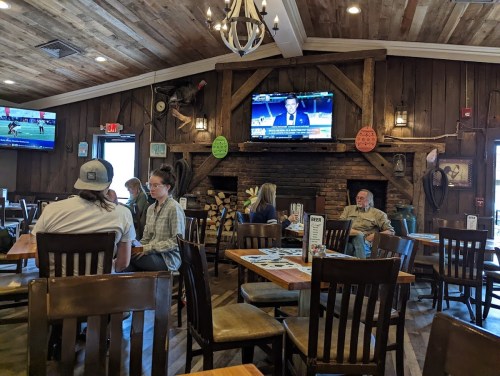 A cozy restaurant interior with wooden decor, tables occupied by diners, and a TV showing news in the background.