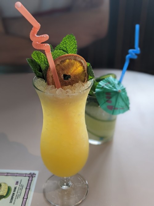 A vibrant yellow cocktail garnished with mint, a dried orange slice, and colorful straws, next to a green drink with an umbrella.