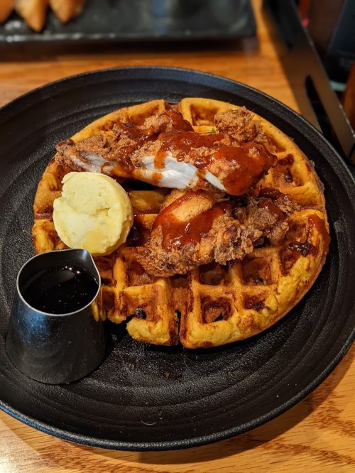 A golden waffle topped with fried chicken, drizzled with sauce, and served with a pat of butter and a small syrup cup.