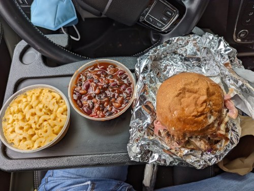 A close-up of a sandwich wrapped in foil, a side of macaroni and cheese, and a container of baked beans on a car seat.