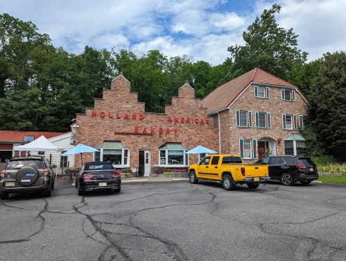 A brick building with "Holland American Bakery" sign, surrounded by cars and outdoor seating under blue umbrellas.