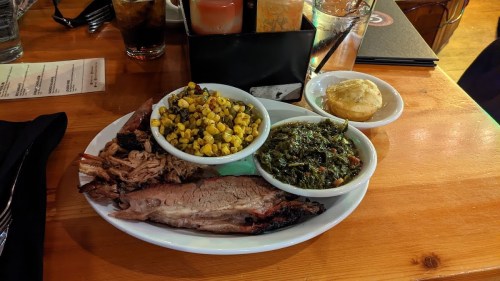 A plate of barbecue brisket, pulled pork, corn, collard greens, and a biscuit on a wooden table.