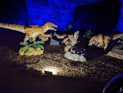 Three dinosaur models in a dimly lit cave, with blue lighting highlighting their features and surrounding rocks.