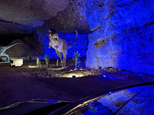 A dinosaur statue illuminated in blue light inside a cave, with plants and a glowing cube nearby.