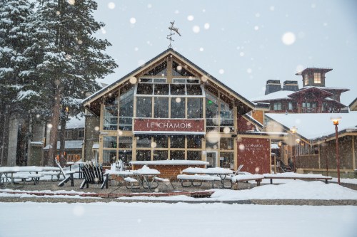 A cozy lodge named "Le Chamois" surrounded by snow, with outdoor seating and snowflakes falling gently.