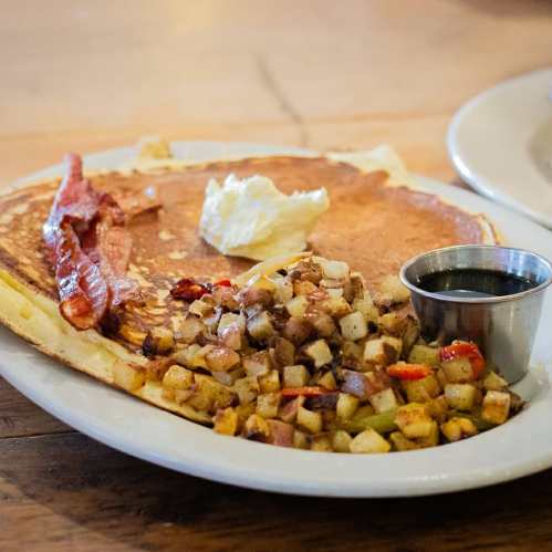 A plate with a large pancake topped with butter, crispy bacon, and a side of seasoned diced potatoes with syrup.