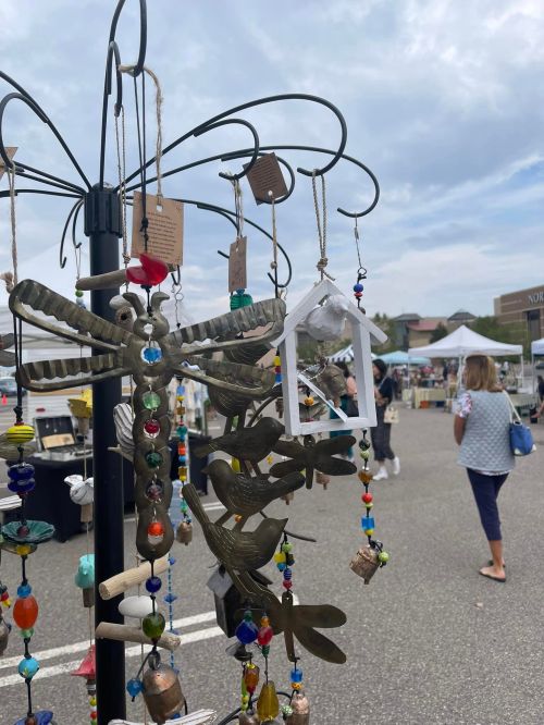 Colorful handmade decorations hang from a display at an outdoor market, with shoppers browsing in the background.